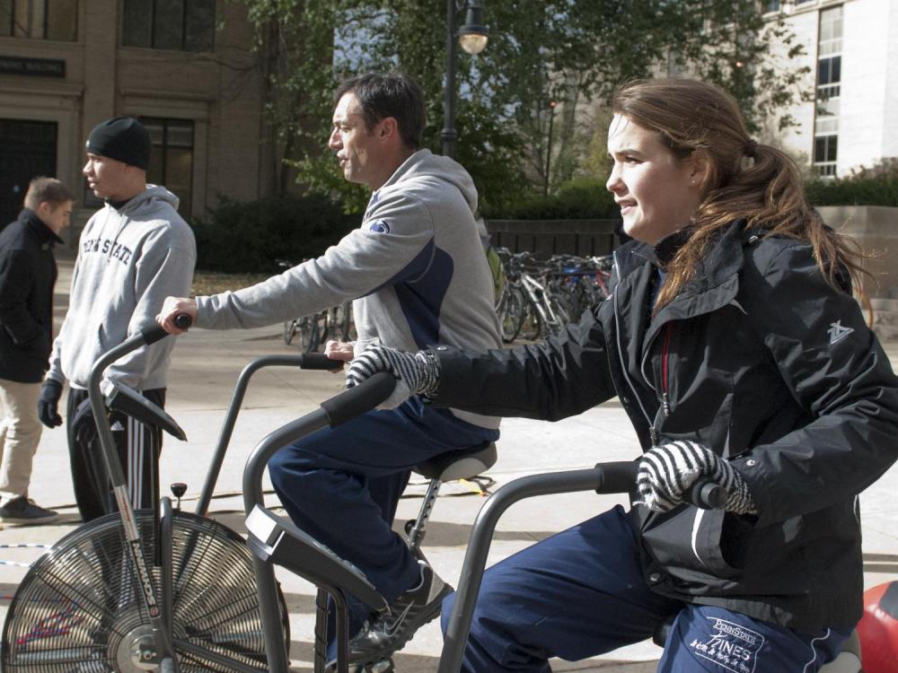 A man and a woman ride stationary bicycles outdoors on University Park campus.