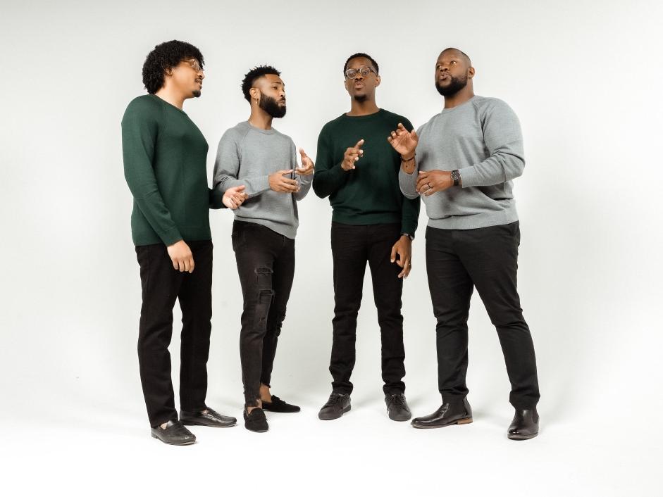 Four men of color stand in a semi-circle and sing in unison.
