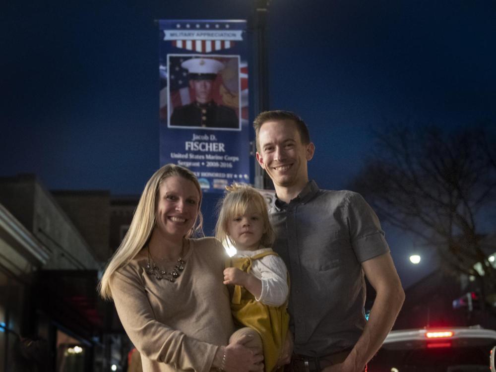 Marine Corps Sgt. Jacob Fischer posing with his wife and daughter in front of his Military Appreciation Week banner in downtown State College.