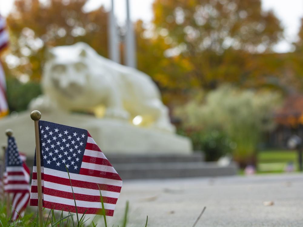 Photo of U.S. flags planted in the grass, with the Nittany Lion statue in the background