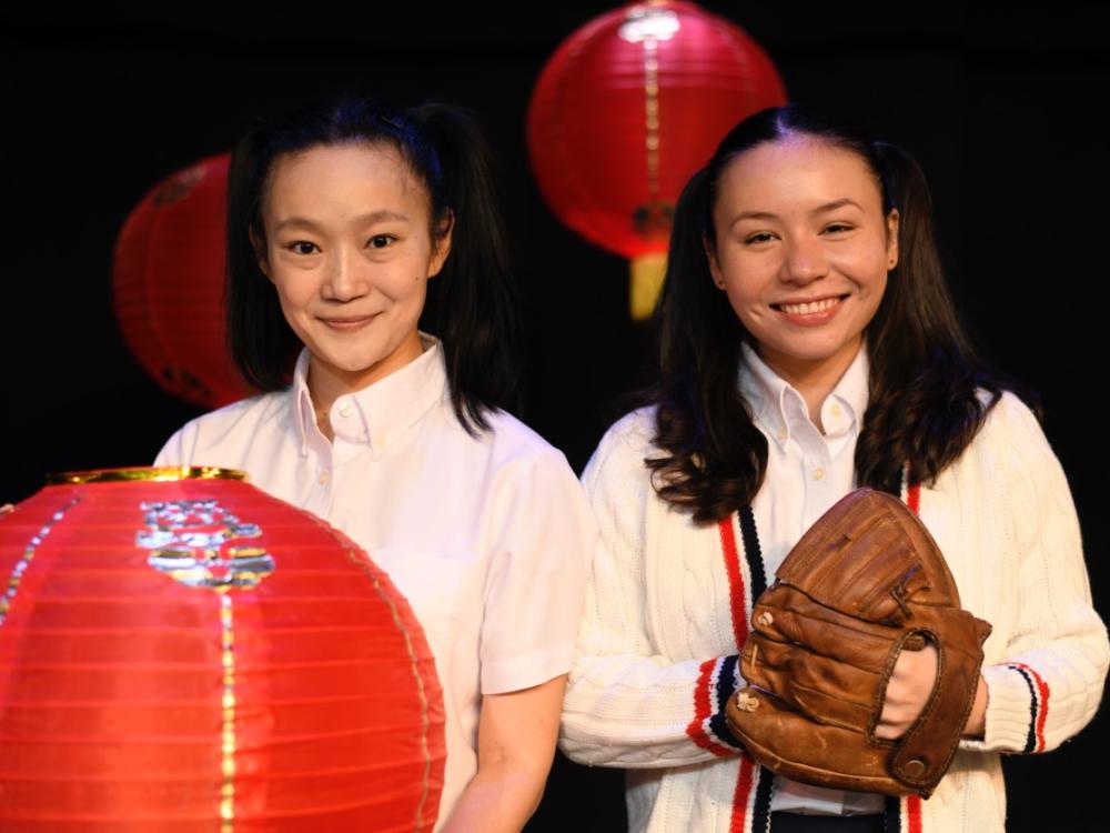 Two girls of Chinese heritage stand shoulder to shoulder, one holding a lantern, the other holding a baseballl glove.