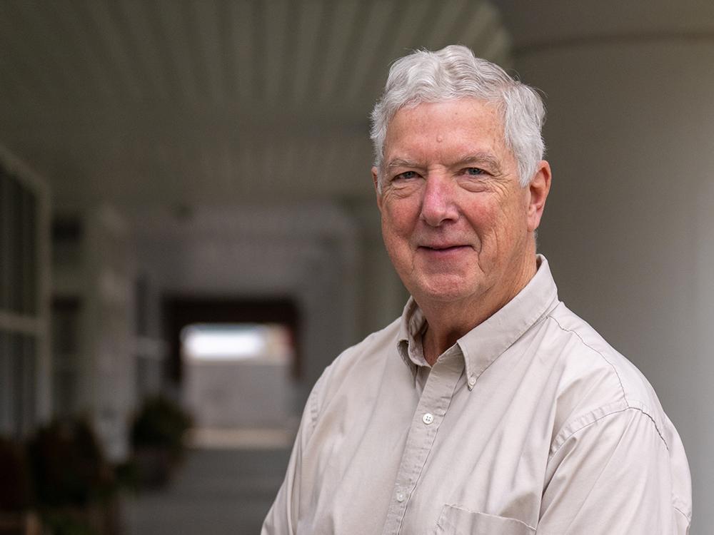 White-haired man in oatmeal-colored oxford stands on the porch of large white building with windows.