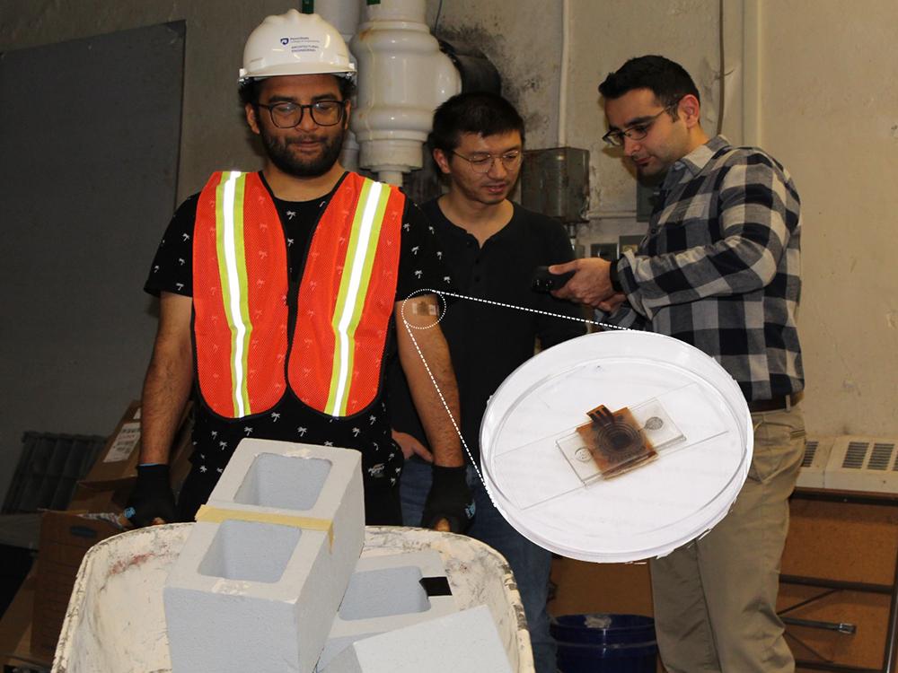 A person in a hard hat and orange vest wears a patch on his arm, which is shown enlarged in the foreground of the image. Two people stand behind the first one.