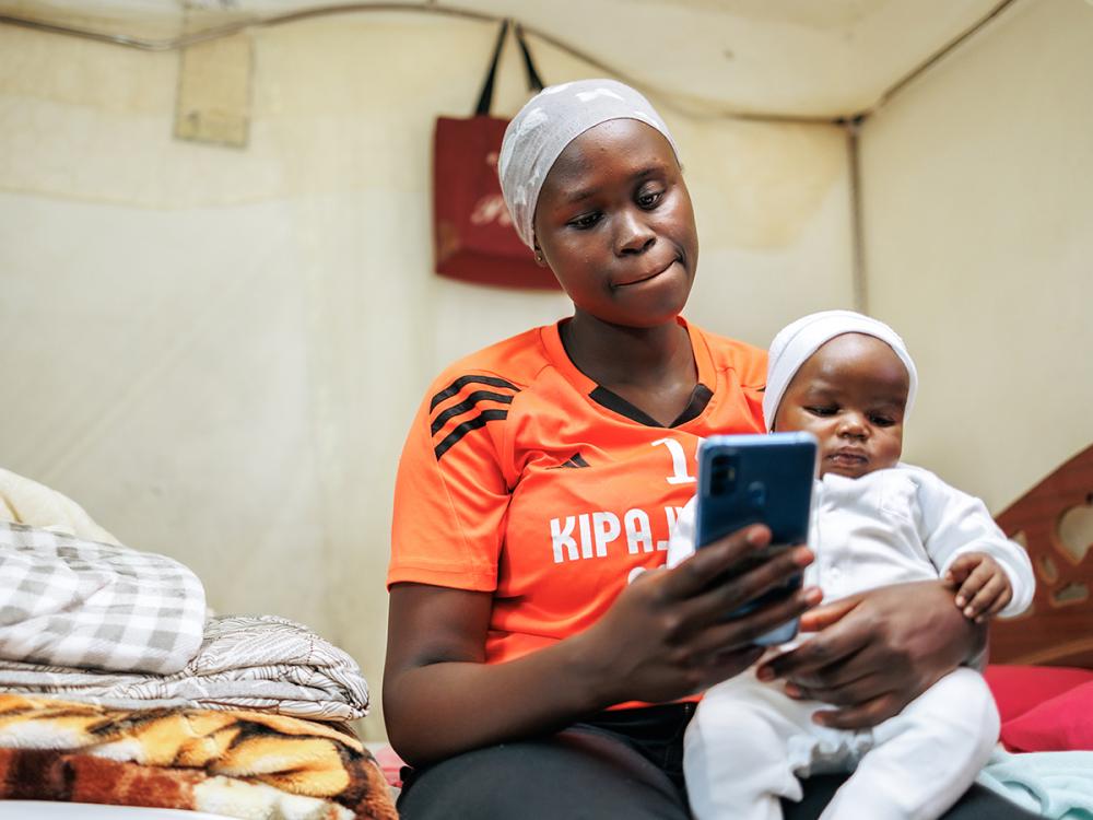 A black woman in an orange shirt looks at her phone while holding a newborn baby.