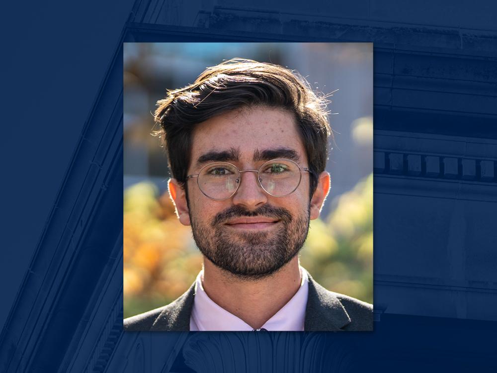 Man with dark hair, beard and glasses wears business suit and smiles in front of outdoor backdrop.