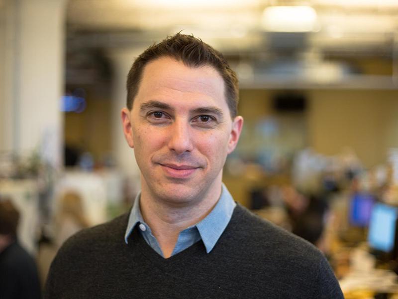 Portrait of Todd Zwillich, smiling, dark haired with collard shirt and sweater