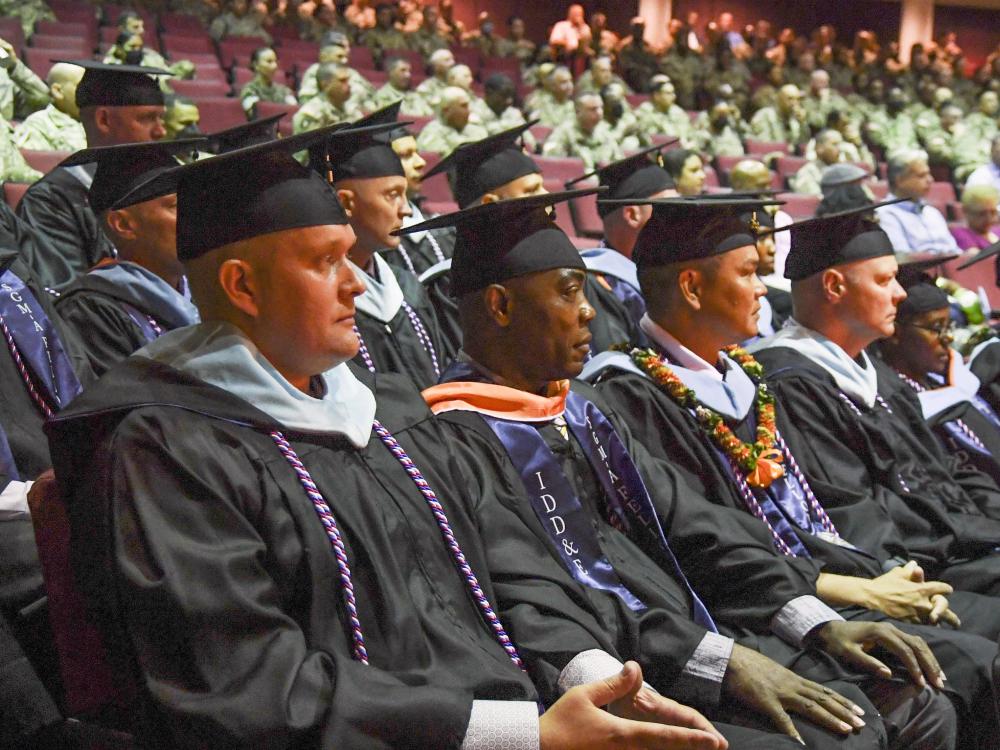 Soldiers wearing graduation caps and gowns and military cords around their necks sit in an auditorium