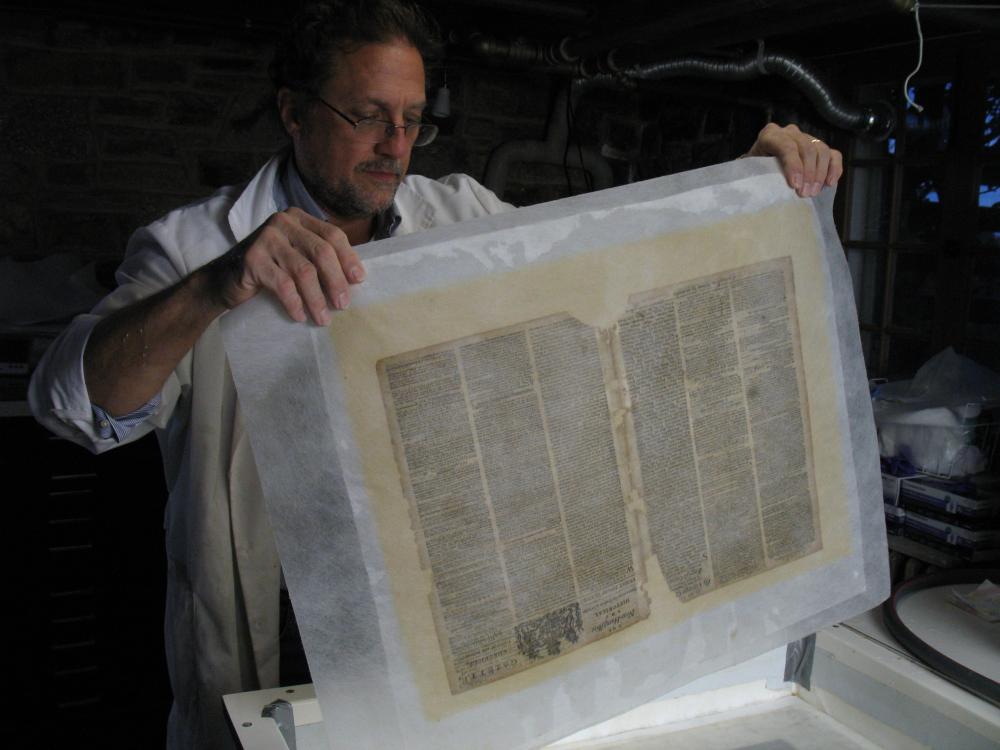 man holding large sheets of sheer paper encasing two pages of old printed newspaper