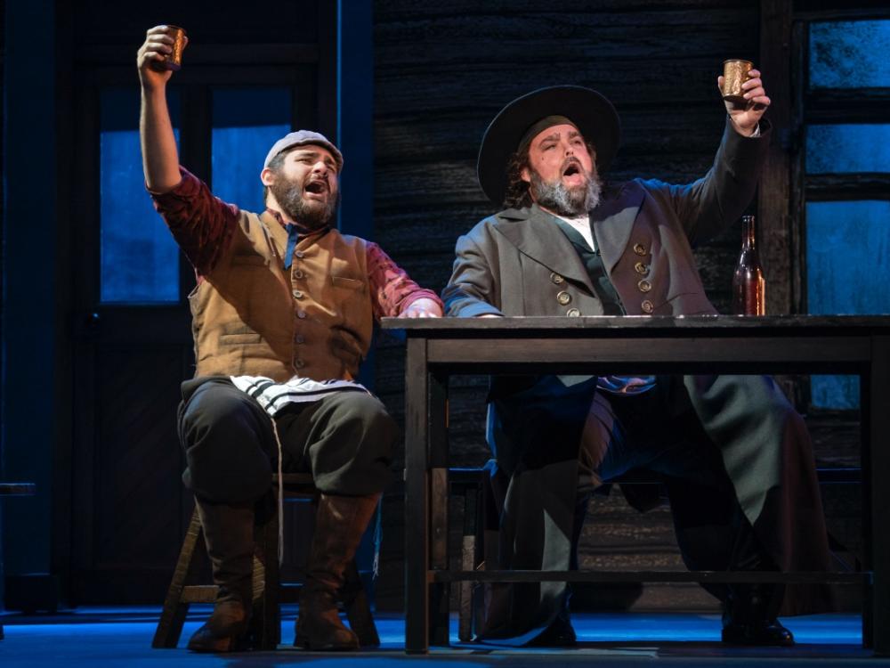 Two husky-figured Jewish men sit at a wooden table and lift their drinks in a toast.