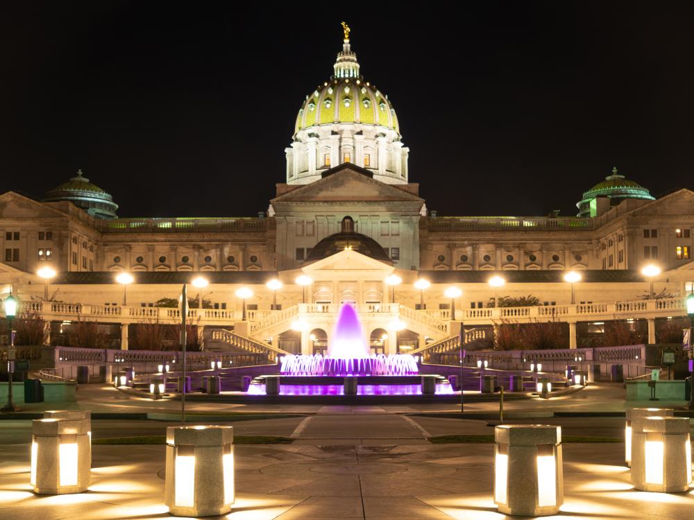 Exterior view of the Pennsylvania State Capitol Building at night with lights and a fountain illuminated in a purple light