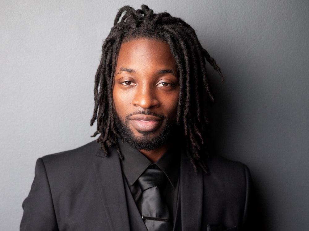 A Black man wearing dreadlocks looks pleasantly chill at the photographer.