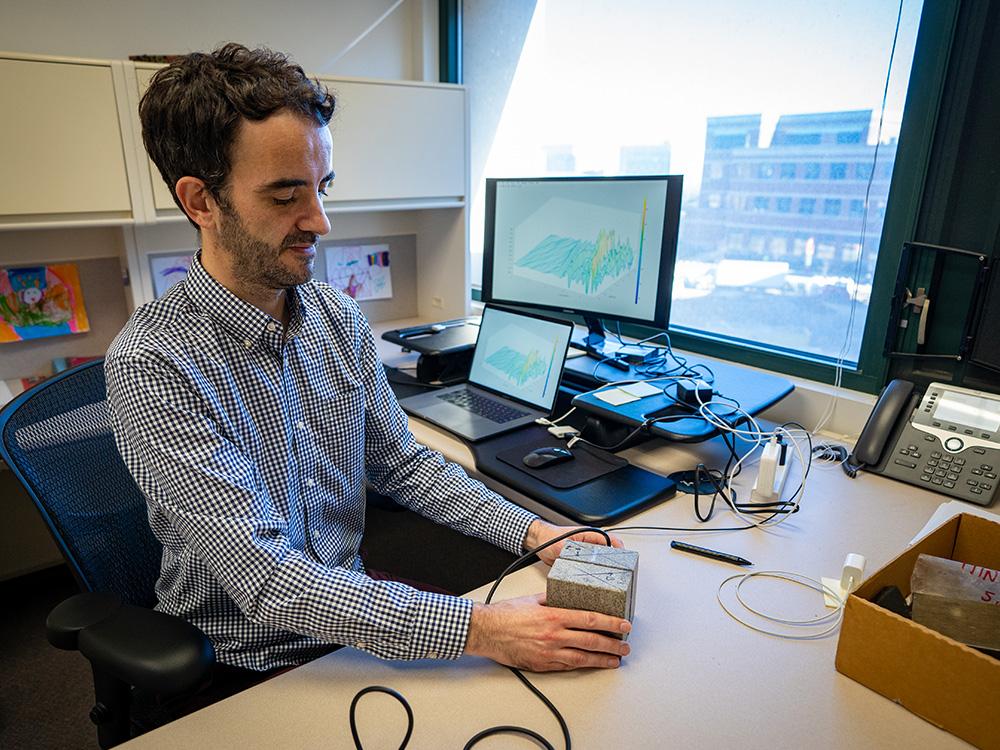 An individual sits at a desk holding two rocks attached to lab instruments.