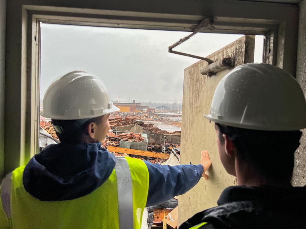 Two students wearing hard hats look through a door at the damage from a tornado.