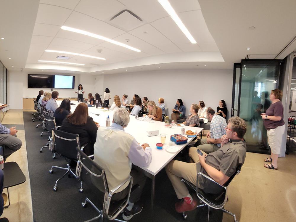 Anjali Gopalakrishnan makes a presentation at the front of a long room to colleagues who are sitting around the rectangular table at Perkins Eastman. 