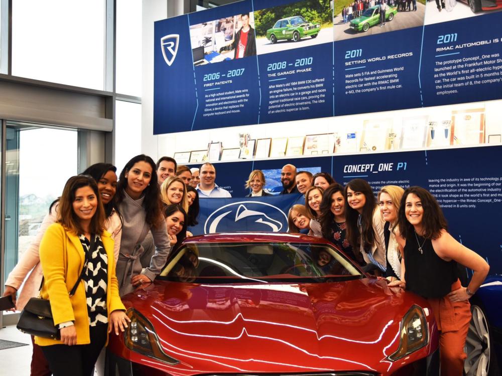 A group of people pose with a Penn State flag in front of a car.