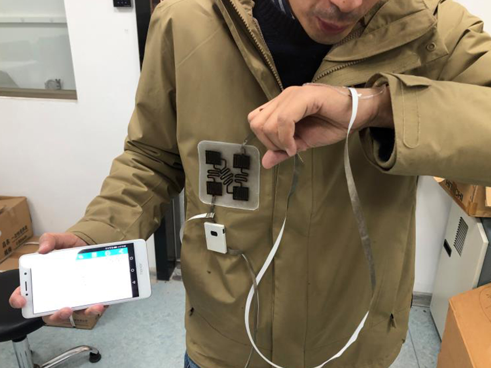 A person in a tan jacket breathes onto their wrist. A wire connects the wrist sensor to a monitor on the jacket. The person holds a phone in their hand, showing data is collected.  