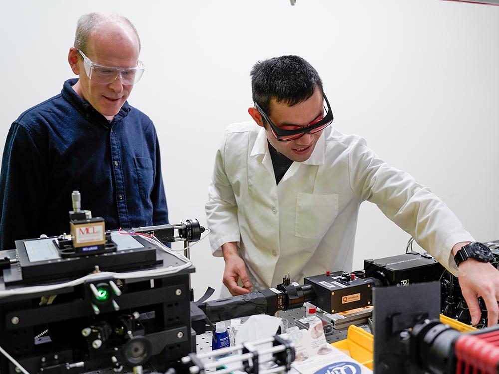 Two individuals wearing lab glasses adjust a microscope in a lab.