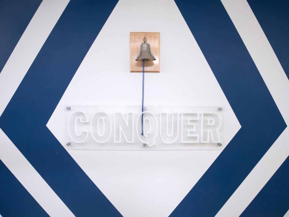 Photo shows a bell that pediatric cancer patients ring during their final cancer treatments at Penn State Health Children’s Hospital. The bell is places above a sign that says, “Conquer.”