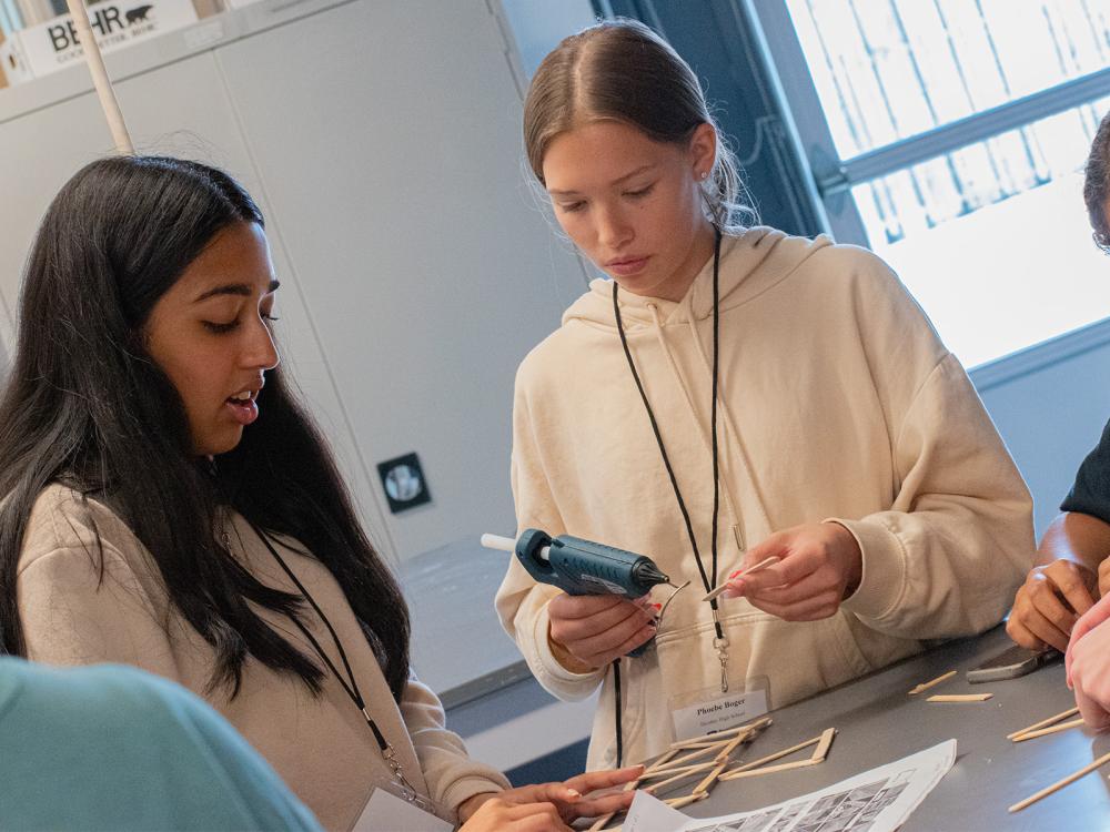 Photo of two students looking at instructions for building a bridge from popsicle sticks. One student holds a hot glue gun.