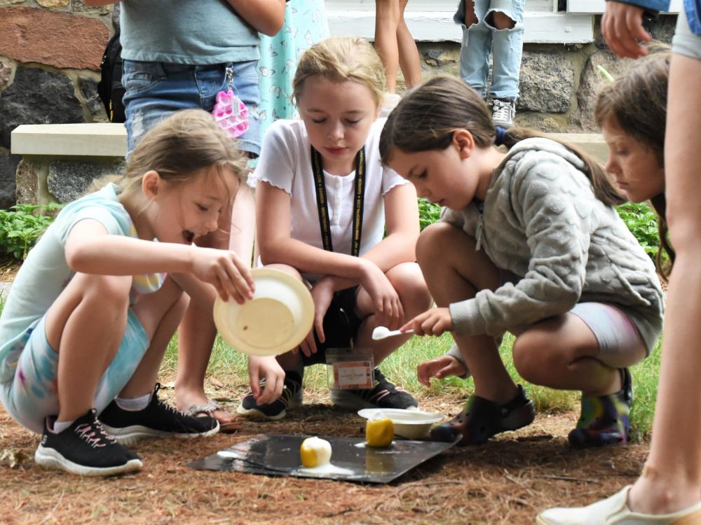 Three girls lean over a food-based science experiment outdoors at Penn State Behrend.