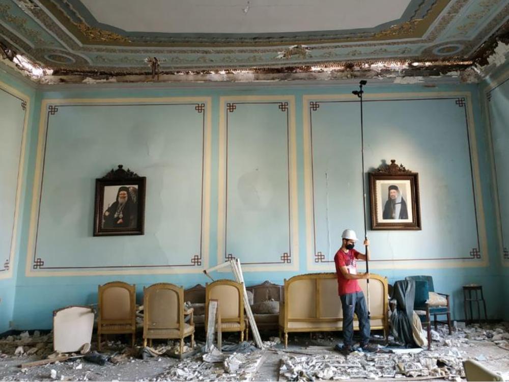 A person wearing a hard hat positions a camera on a tall stick in a blue room with clear damage on the floor and ceiling.