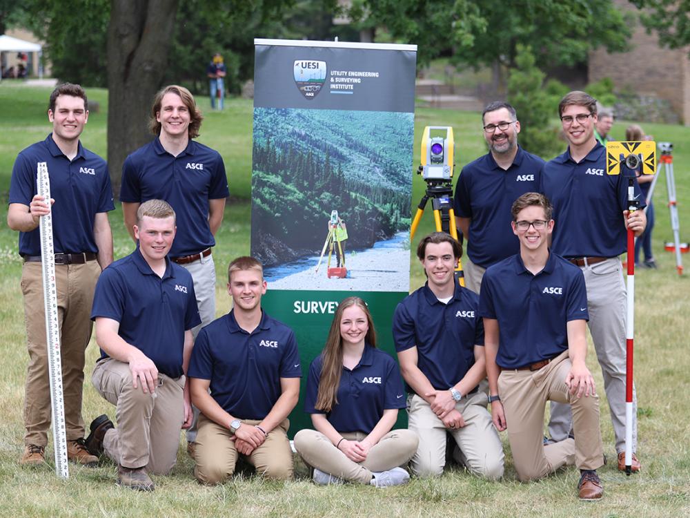 A group of students wearing navy blue polo shirts poses outdoors with surveying equipment. 