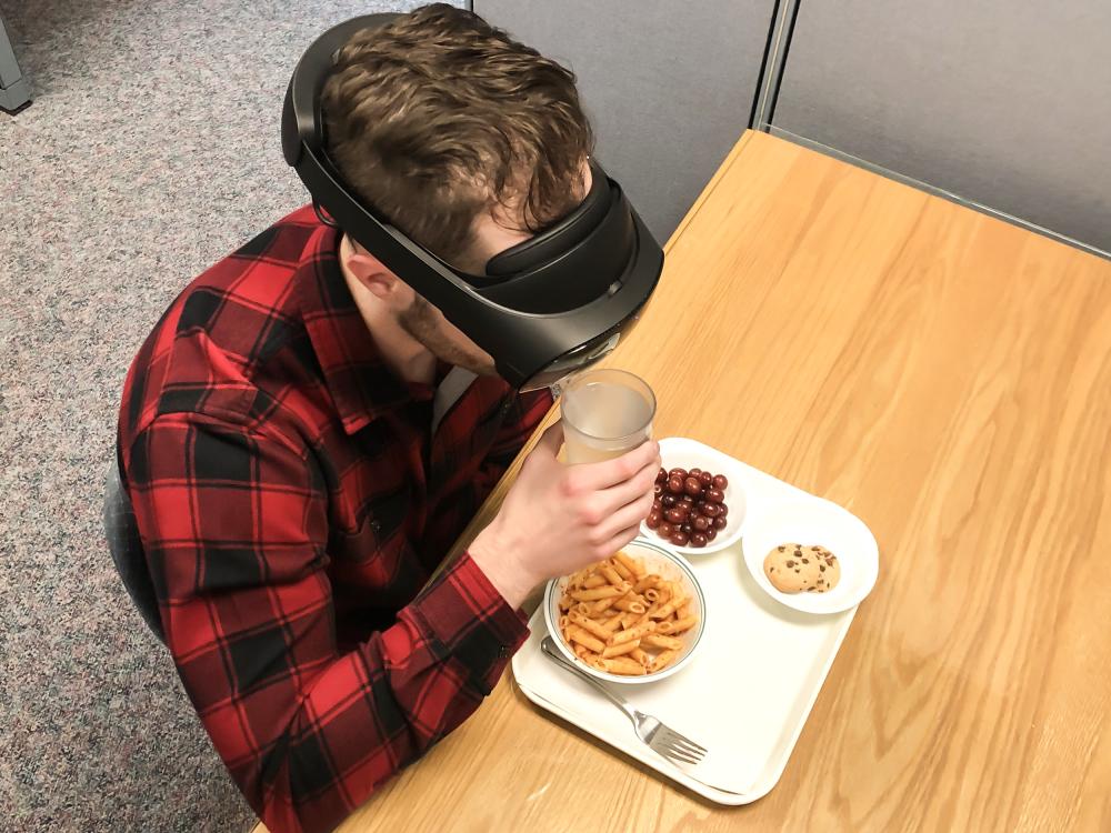 Man sits in cubicle wearing a virtual-reality headset with a meal in front of him. He is drinking from a glass.