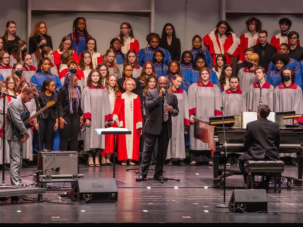 A choir comprised of a diverse group of people stands in a group while a man of color stands in the center and speaks into a microphone.