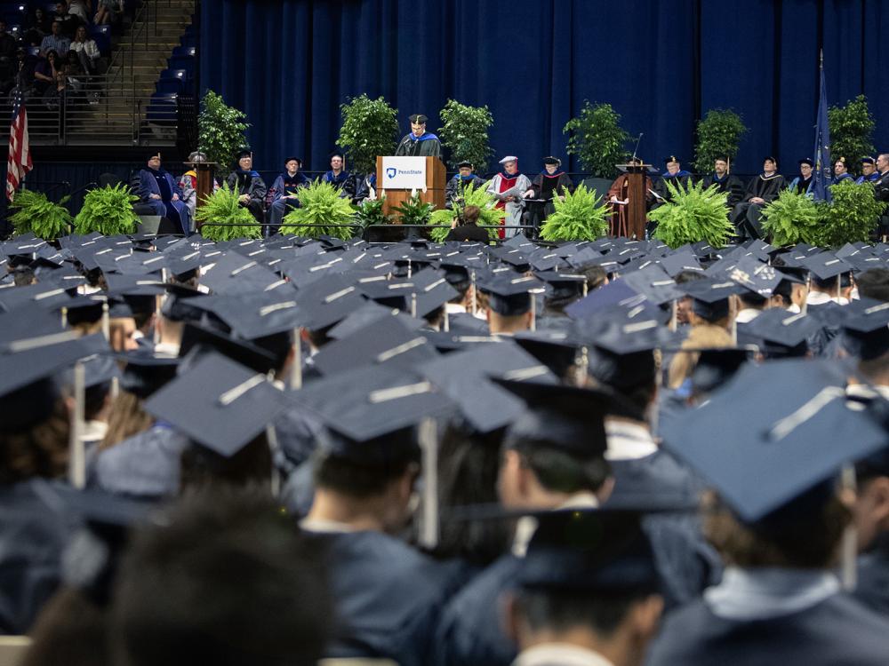 Rows of graduates at commencement in the Bryce Jordan Center for a podium and speaker in the background