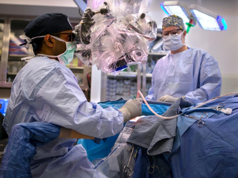 A doctor wearing a surgical gown sits next to an operating table, treating a patient. A nurse wearing a surgical gown stands in the background.