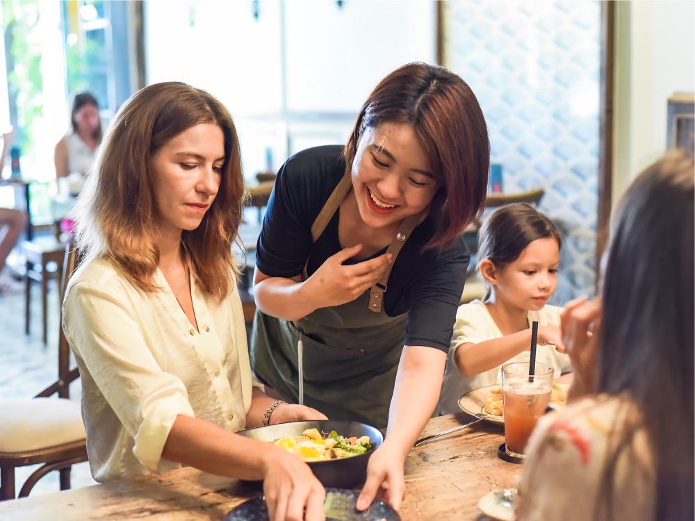 Asian waitress smiles as she leans over a white woman in a busy restaurant