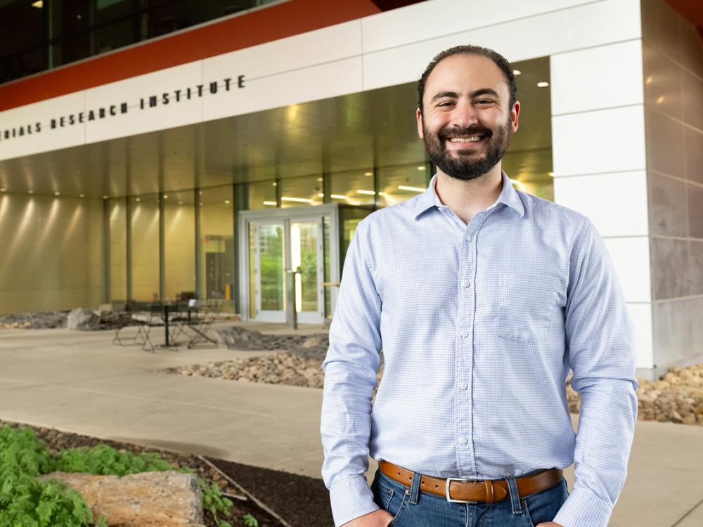 A person in a button-down shirt stands in front of the Materials Research Institute building