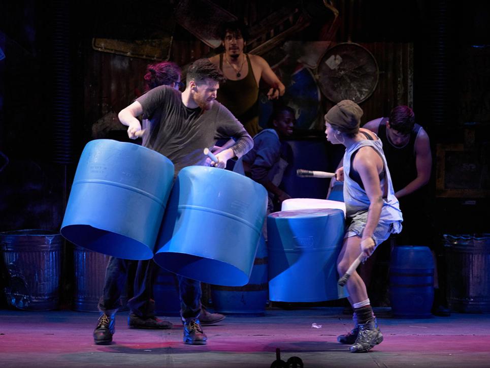 Two performers wear plastic drums around their shoulders while they use sticks to bang on them.