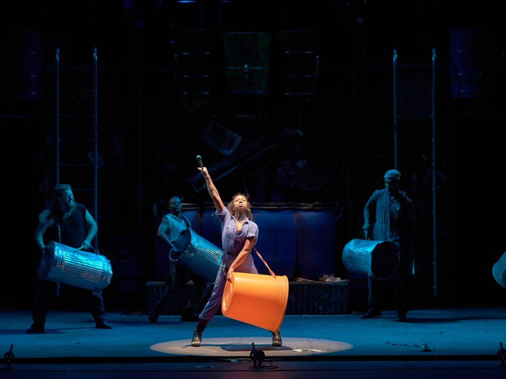 A woman of color wearing a large plastic drum on a strap around her neck and shoulders, looks up and raises a hand holding a drumstick.