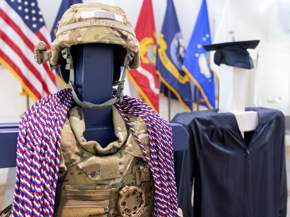 Red, white and blue honor cords are draped over camouflaged military gear next to a blue graduation cap and gown.