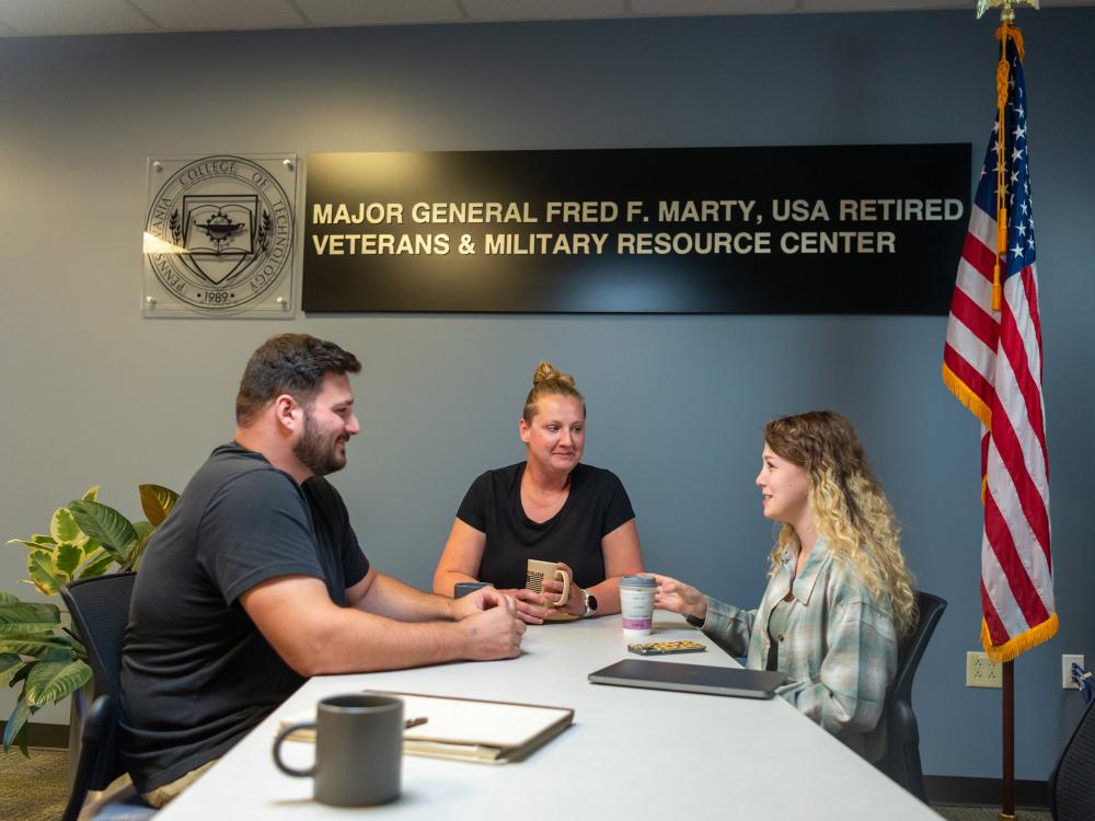 Three people sit at a table for a meeting with a U.S. flag on a stand to the right