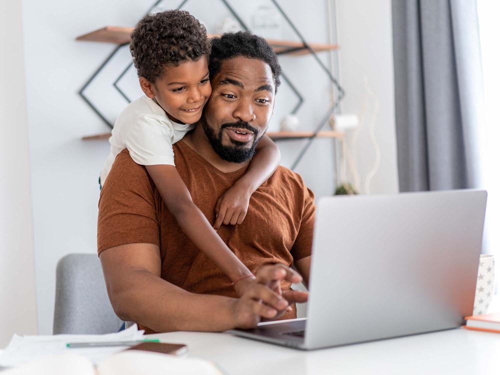 A father working at home on a laptop with his son's arms wrapped around him.