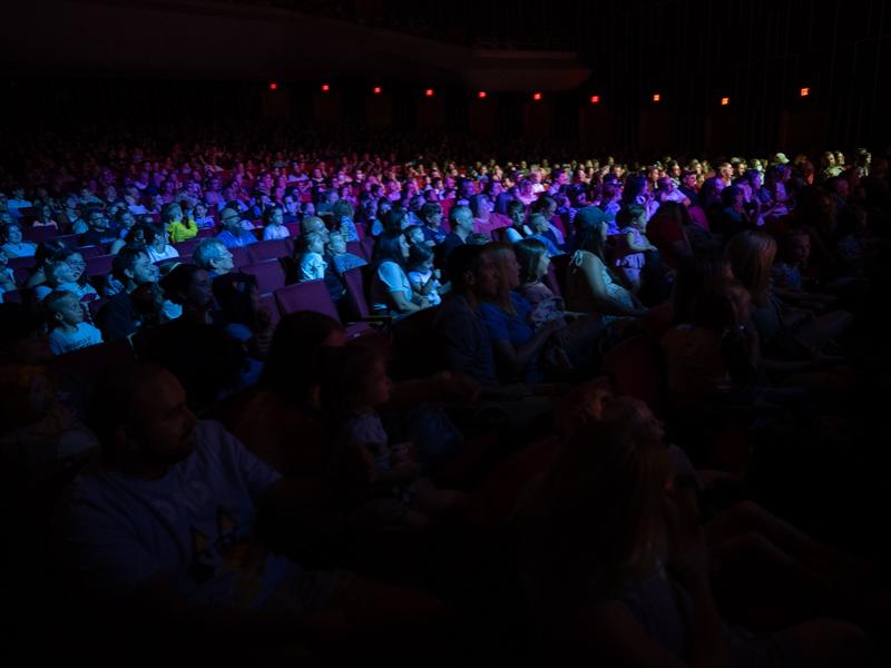 A diverse audience sits in a dimly lit auditorium and watch a performance on the stage.