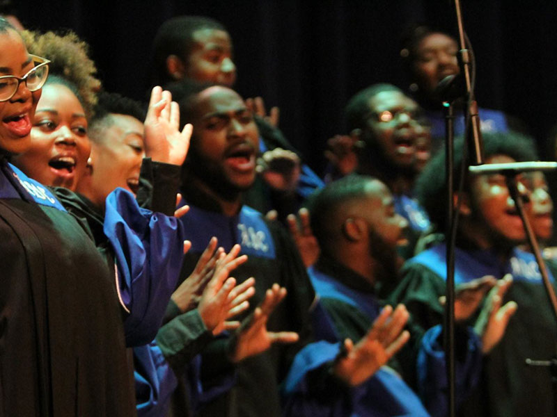 Black artists dressed in matching robes raise their hands while they sing.