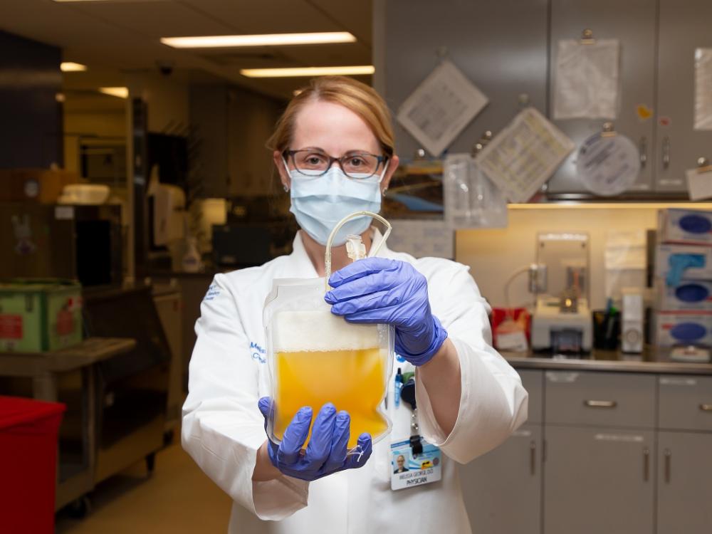 A woman in scrubs and a surgical mask holds a blood bag full of plasma.