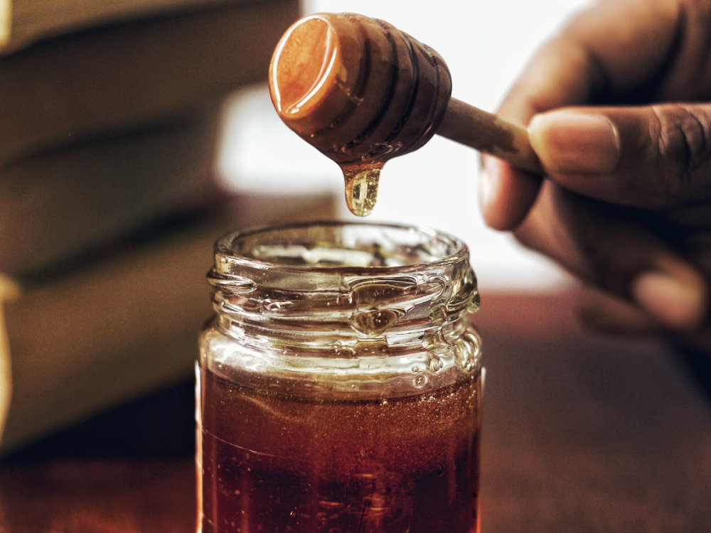 A hand holding a honey dipper over a jar of honey
