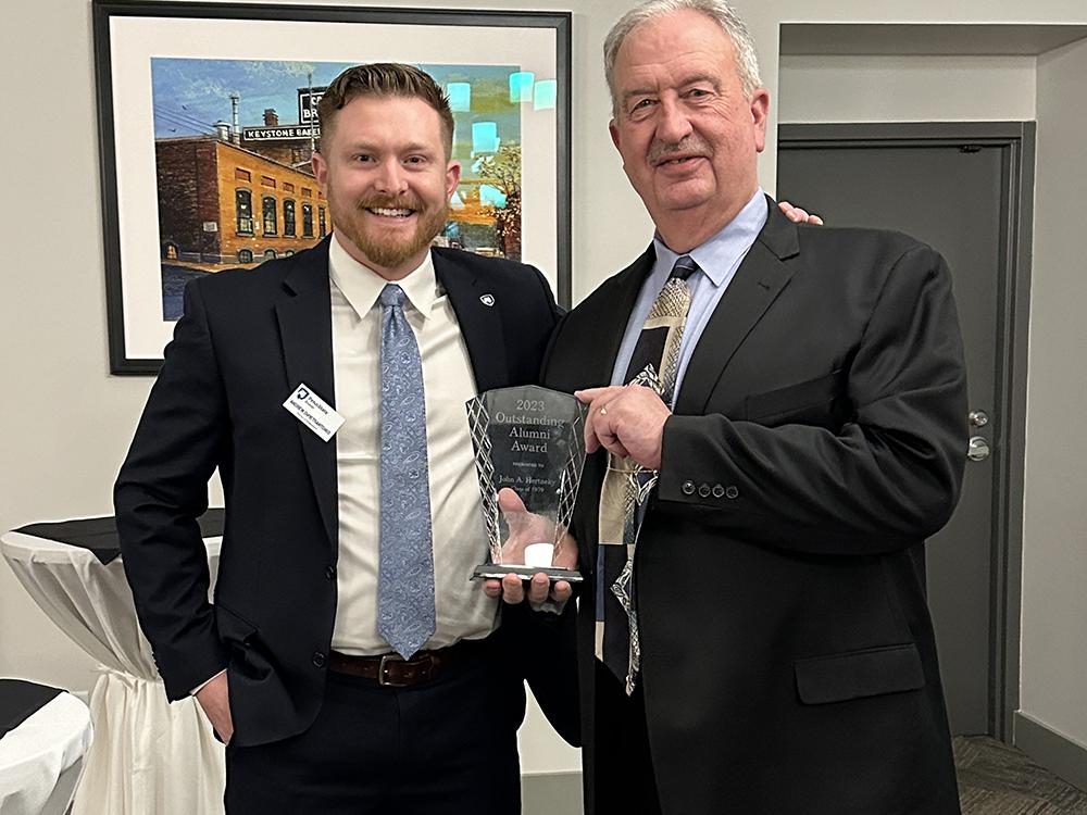 Hertneky stands at the right of the photo holding a glass award handed to him by Andrew DiPietrantonio who stands at left