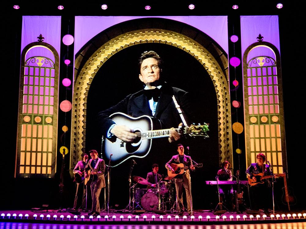 A band of musicians perform on stage while a screen behind them shows footage of Johnny Cash performing.