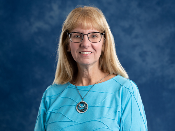 Woman with long blonde hair, glasses and light blue long-sleeved shirt.