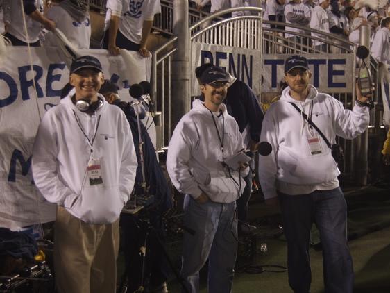 Three men stand on the sidelines at Beaver Stadium in front of the crowd