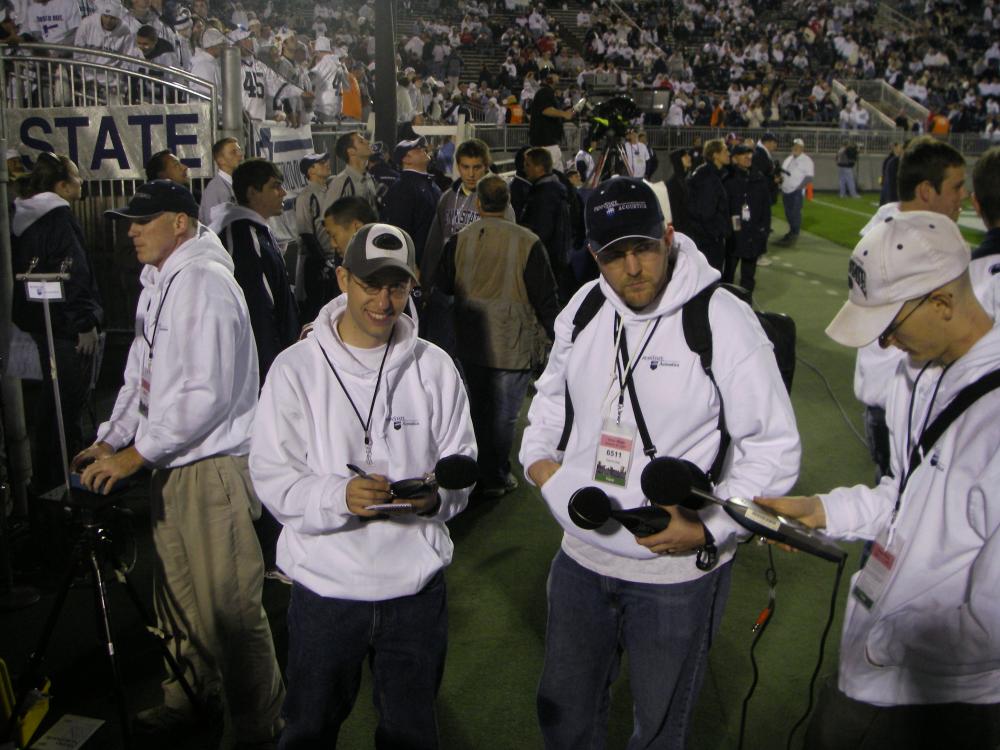 Four men stand with recording equipment and sound measuring tools on the field at Beaver Stadium