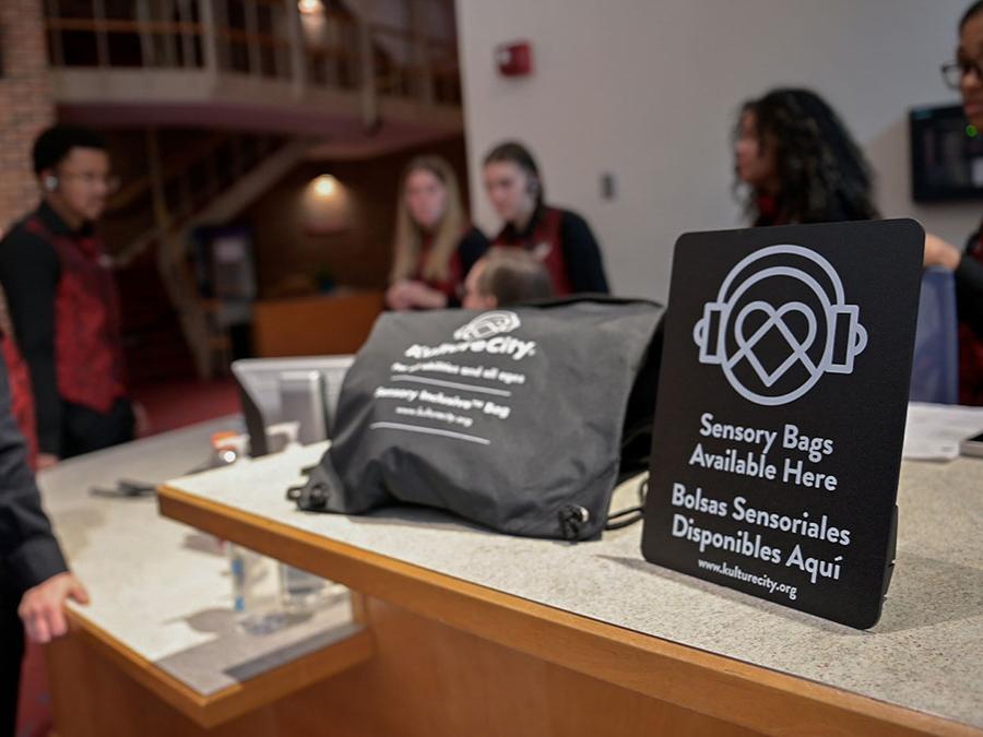 A KultureCity swag bag sits on a counter next to a sign while a group of volunteers stand in the background.