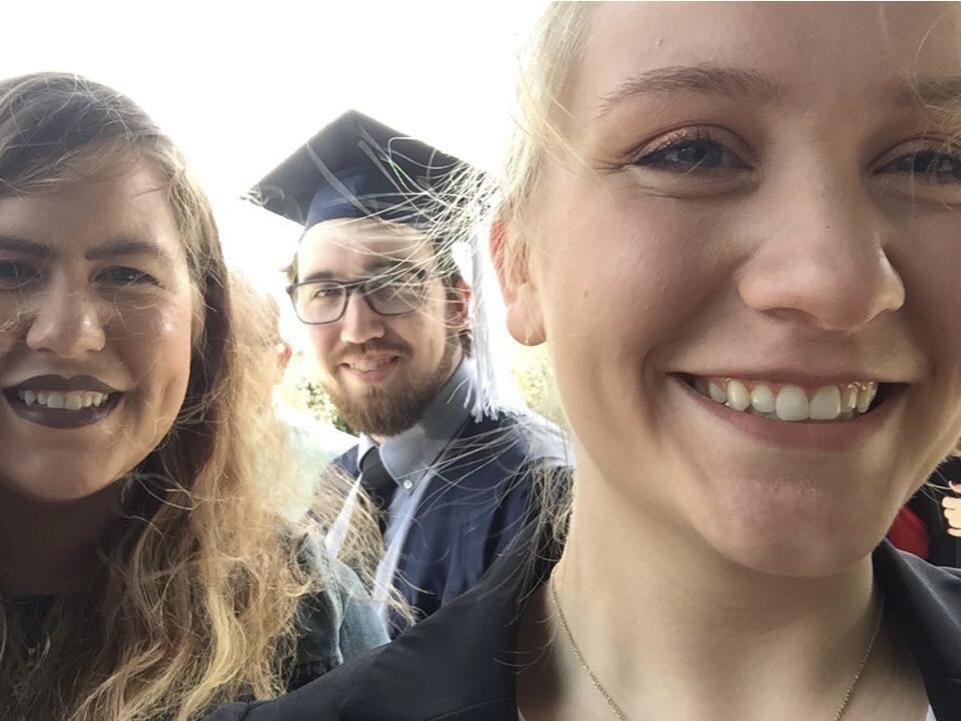 Two white young women and a white young man wearing a blue graduation cap