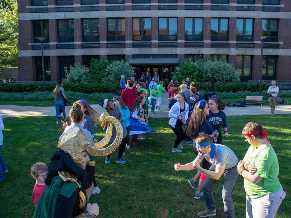 A group of people of all ages wearing loose clothing and costumes stand in two facing rows on a grassy lawn and make dance poses.