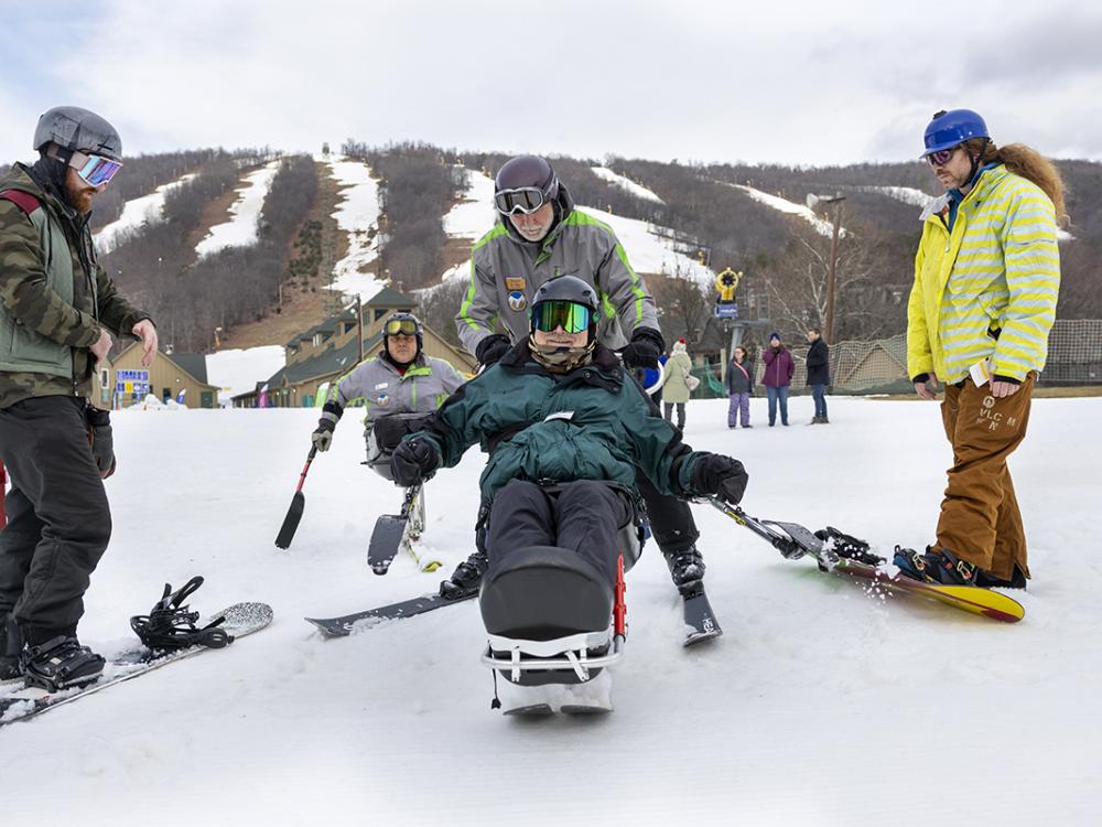 A group of people in parkas riding skis gather at the base of a mountain striped with ski runs. One is seated on a sit-ski, a device that balances his body on a single runner, and his legs are propped in front of him.